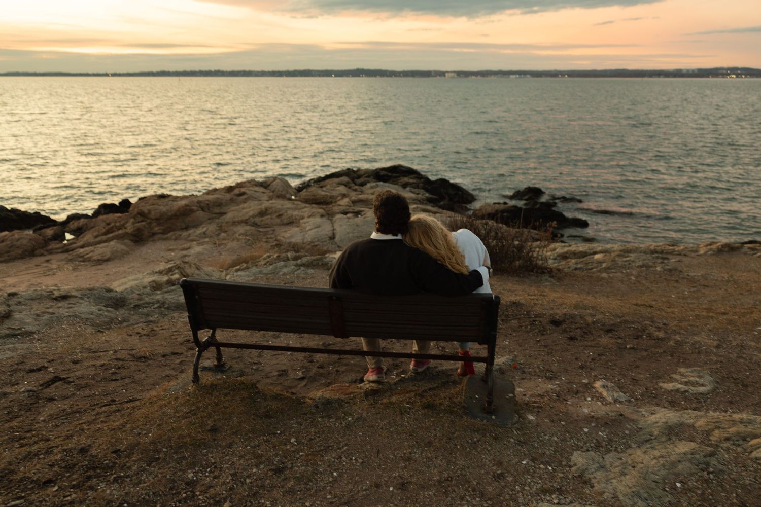 A couple sits together on a bench overlooking the ocean at sunset.