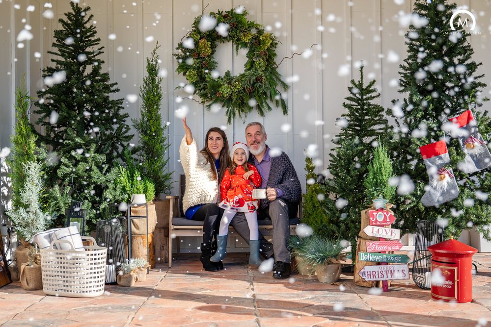 A festive holiday photo booth setup with Christmas trees, white lights, and rustic wooden decor against a white wall.