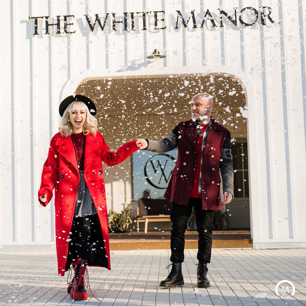 A couple in red winter coats walks together on a snowy day outside a building with white columns.