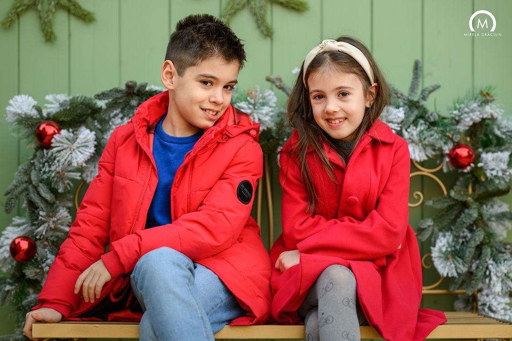 Family wearing red winter coats poses together in front of decorated Christmas tree and string lights.