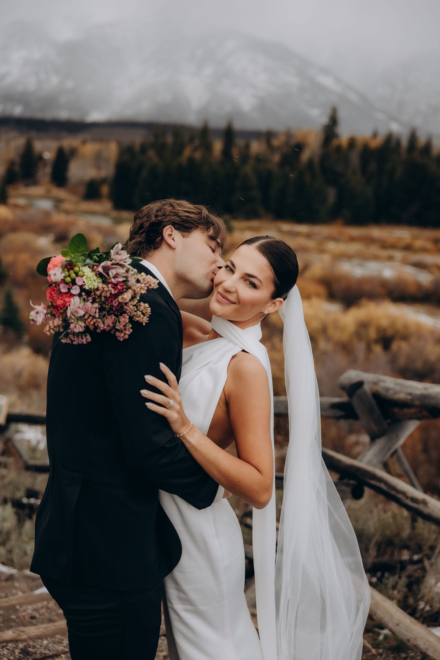 A romantic wedding couple embraces outdoors in a rustic mountain setting with a flowing white veil and autumn colors.