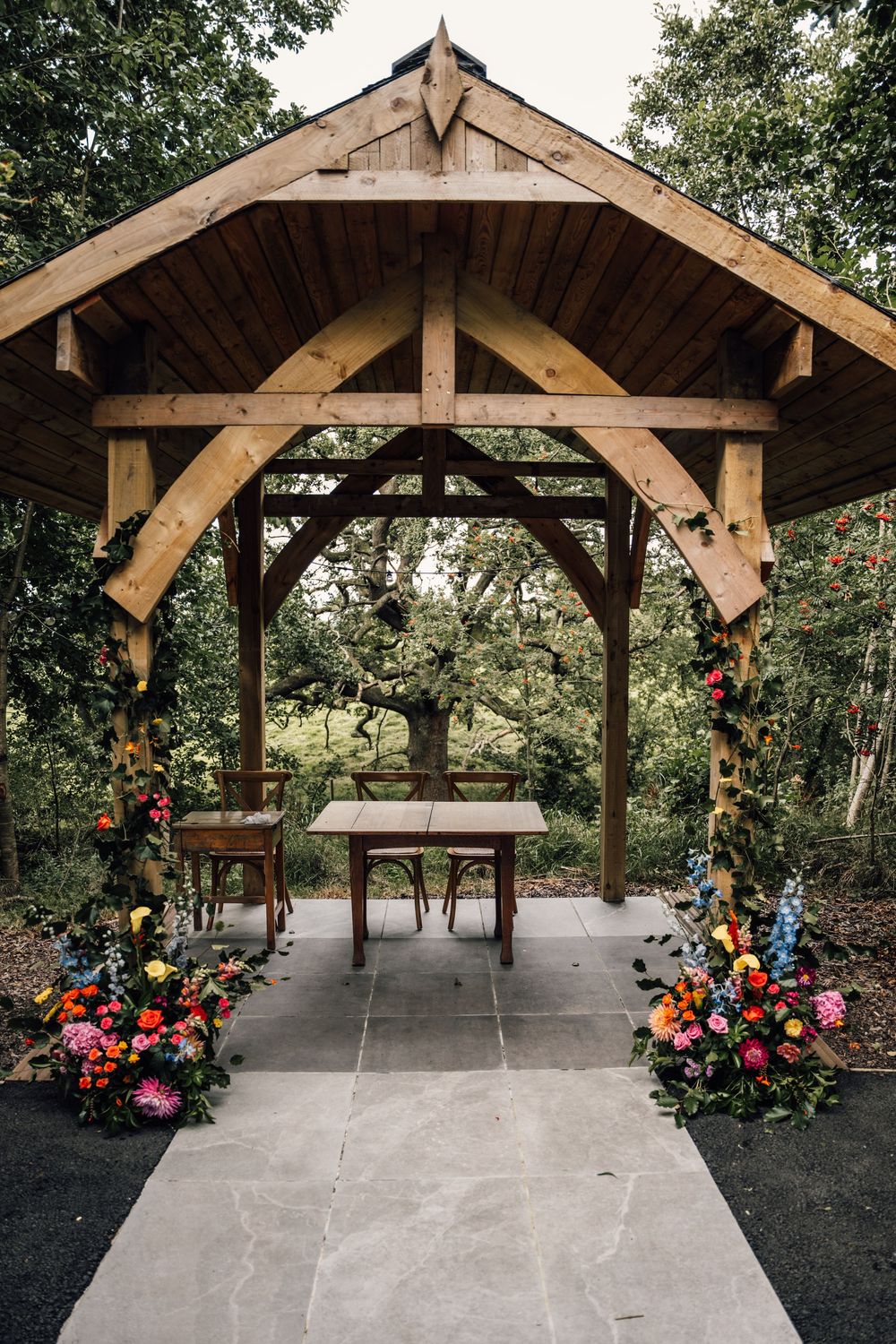 Rustic wooden pavilion decorated with colorful floral arrangements in an outdoor forest setting.