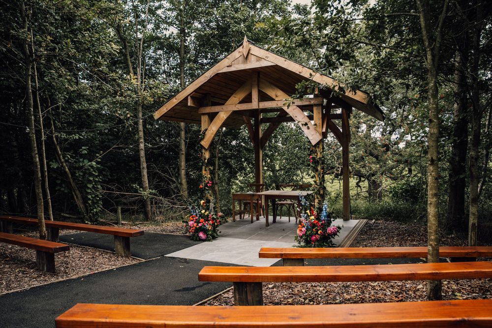 Open-air timber frame pavilion with wooden benches surrounded by trees and natural landscaping.