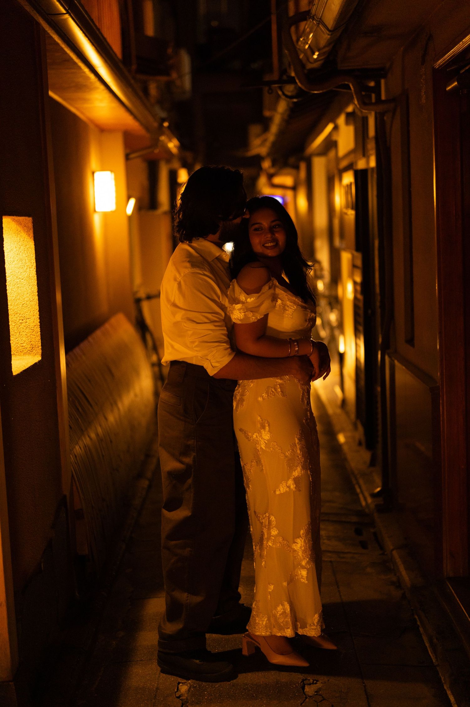 Couple embracing romantically in a warmly lit narrow alley at night, woman in white floral dress.