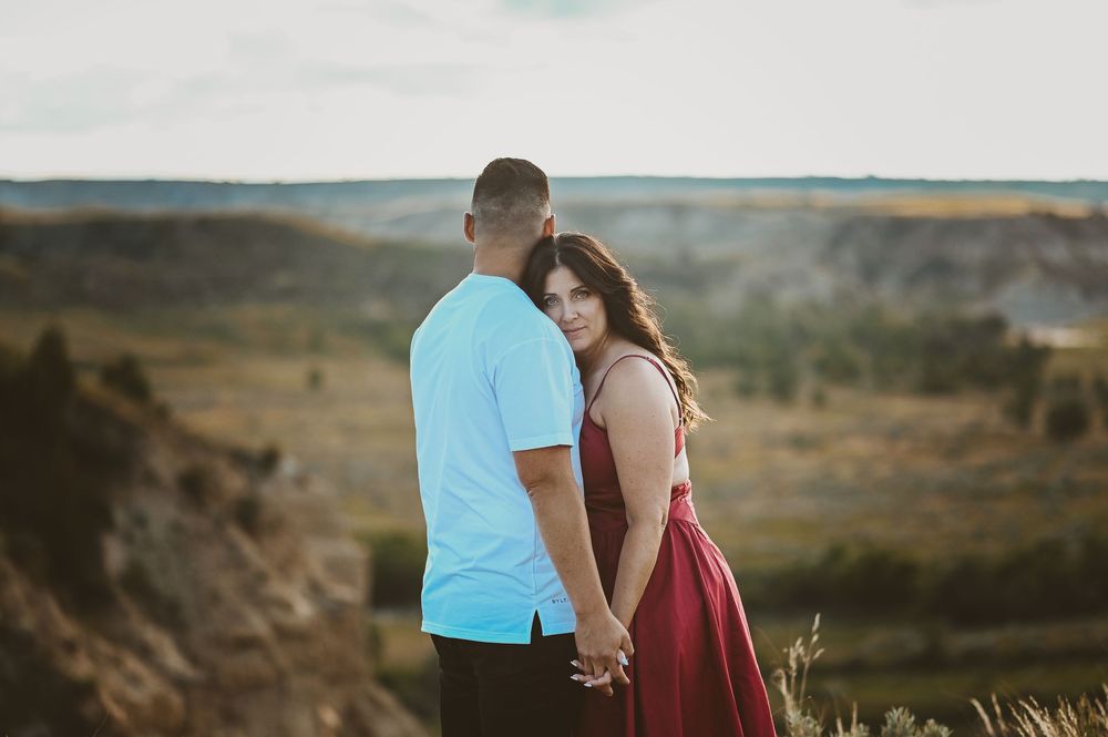 A couple stands close together against a scenic mountain landscape backdrop at sunset.