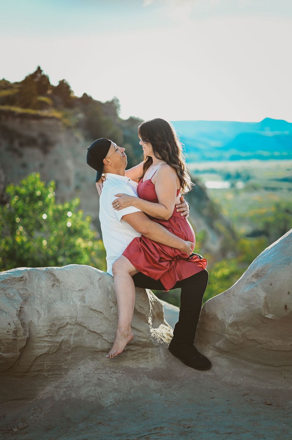 A romantic couple embraces while sitting on rocky cliffs overlooking a scenic mountain landscape at sunset.