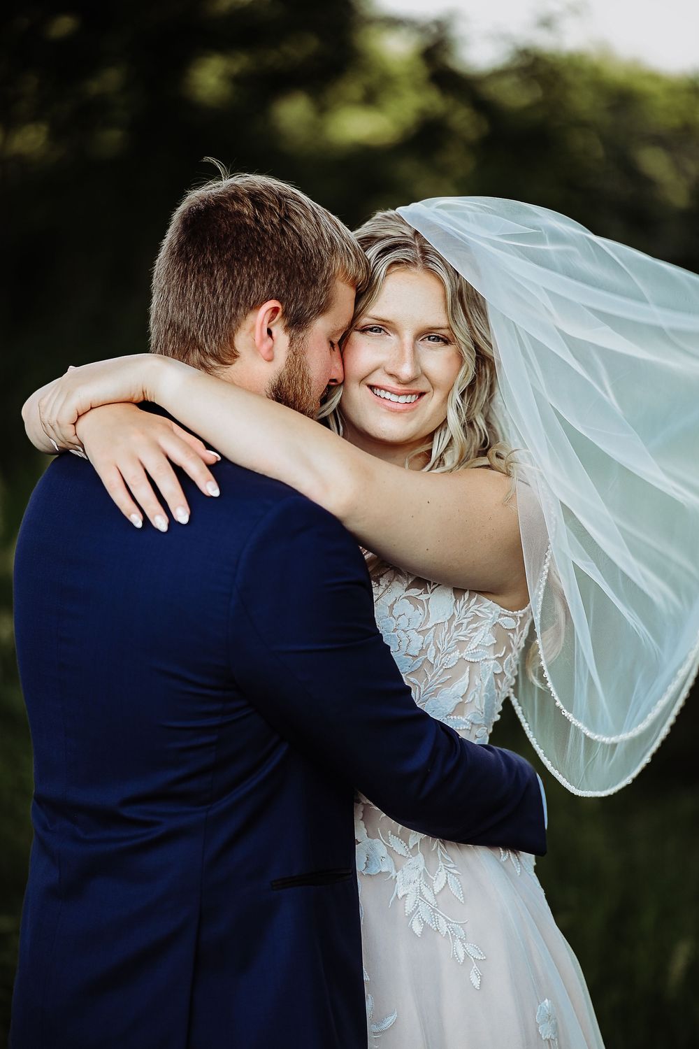 Romantic wedding couple embraces outdoors in natural light with bride wearing flowing veil and lace dress.