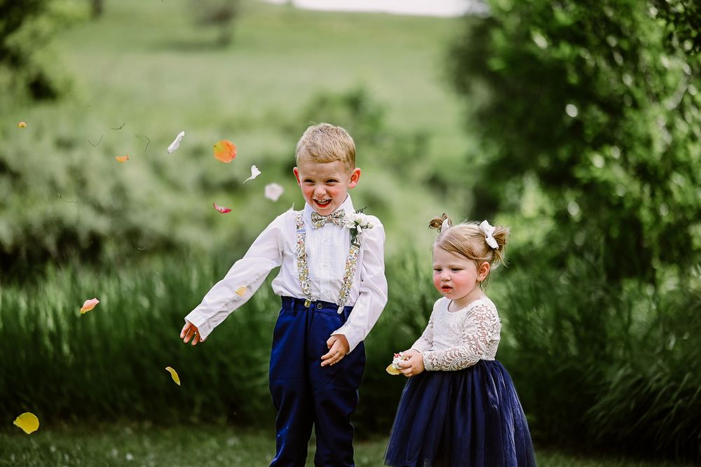 Two young children in formal wear play with falling autumn leaves in a lush green garden setting.