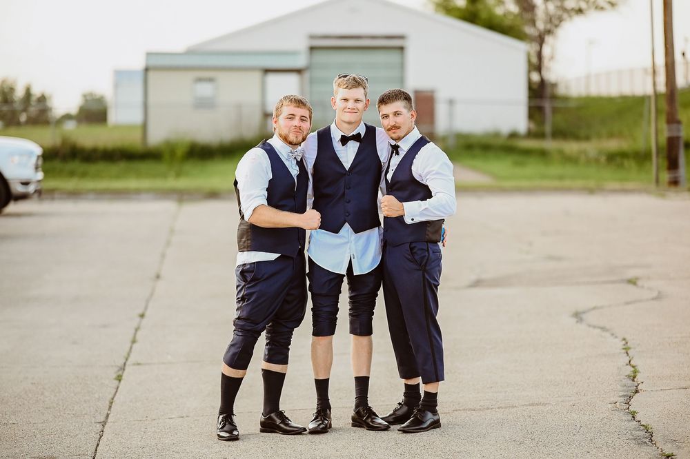 Three young boys in matching navy vests and bow ties stand close together on a concrete path.