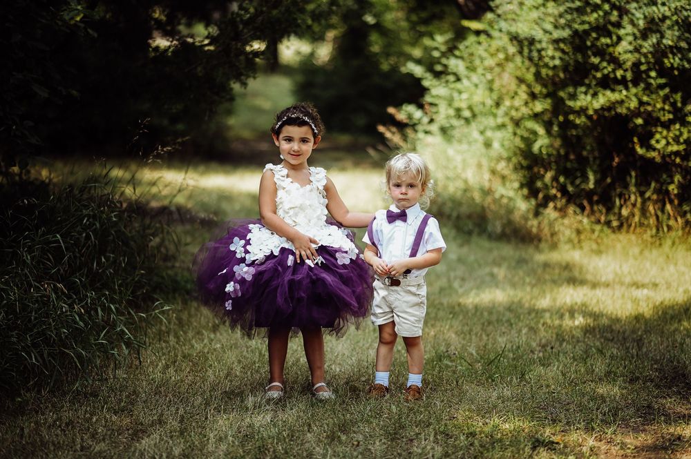 Two children in formal attire walk together on a grassy outdoor path during a sunny day.