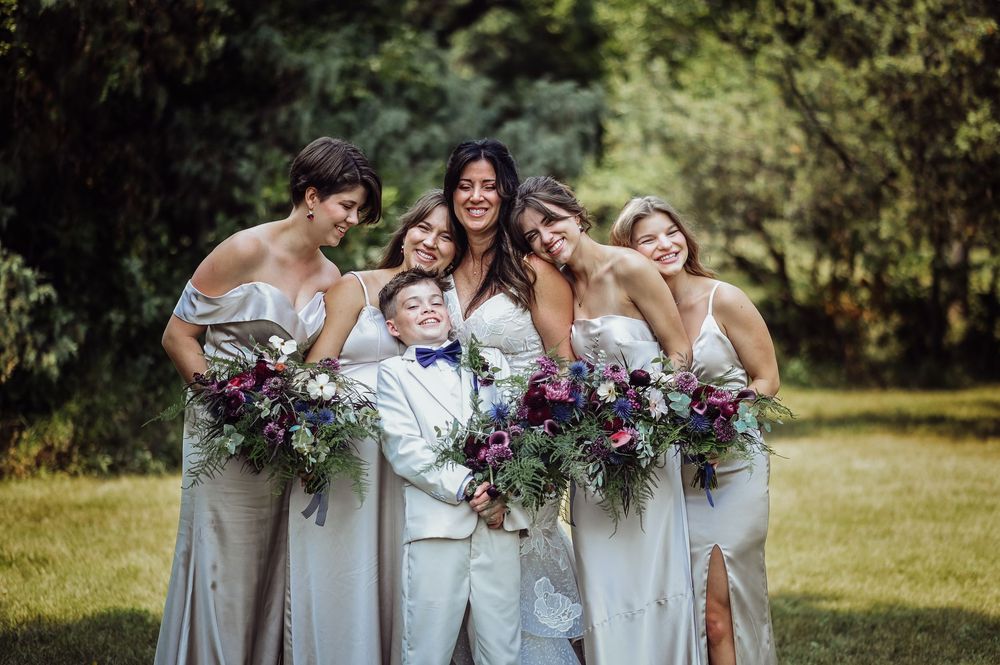 Wedding party in light blue bridesmaid dresses laughs together while holding purple and white bouquets in a garden setting.
