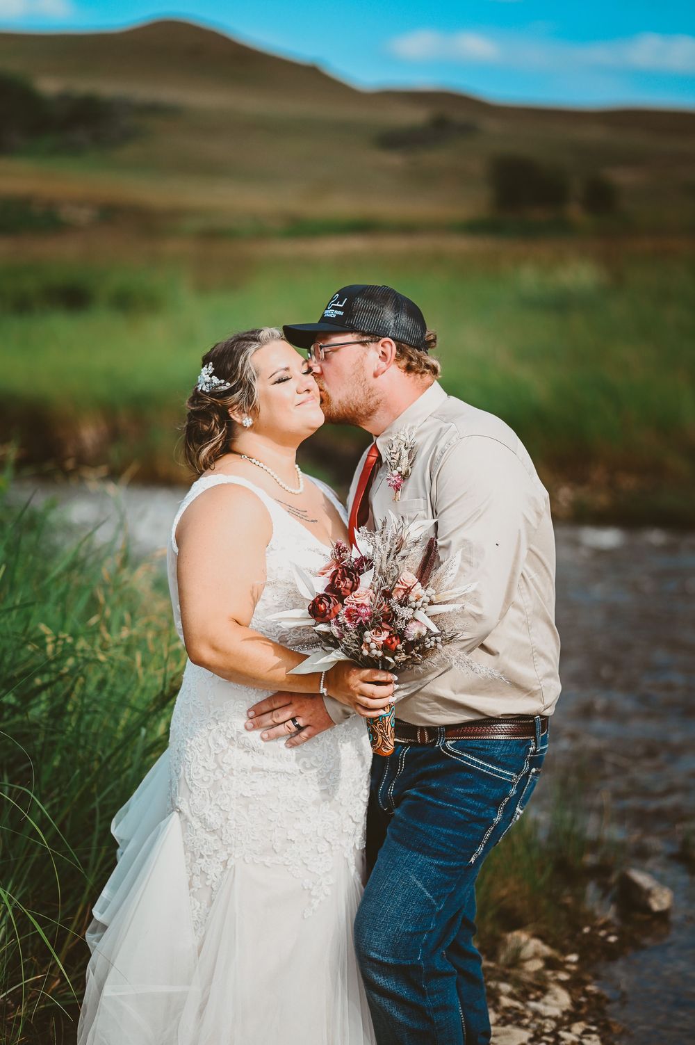 A couple shares an intimate moment by a river with rolling hills in the background during their rustic outdoor wedding.