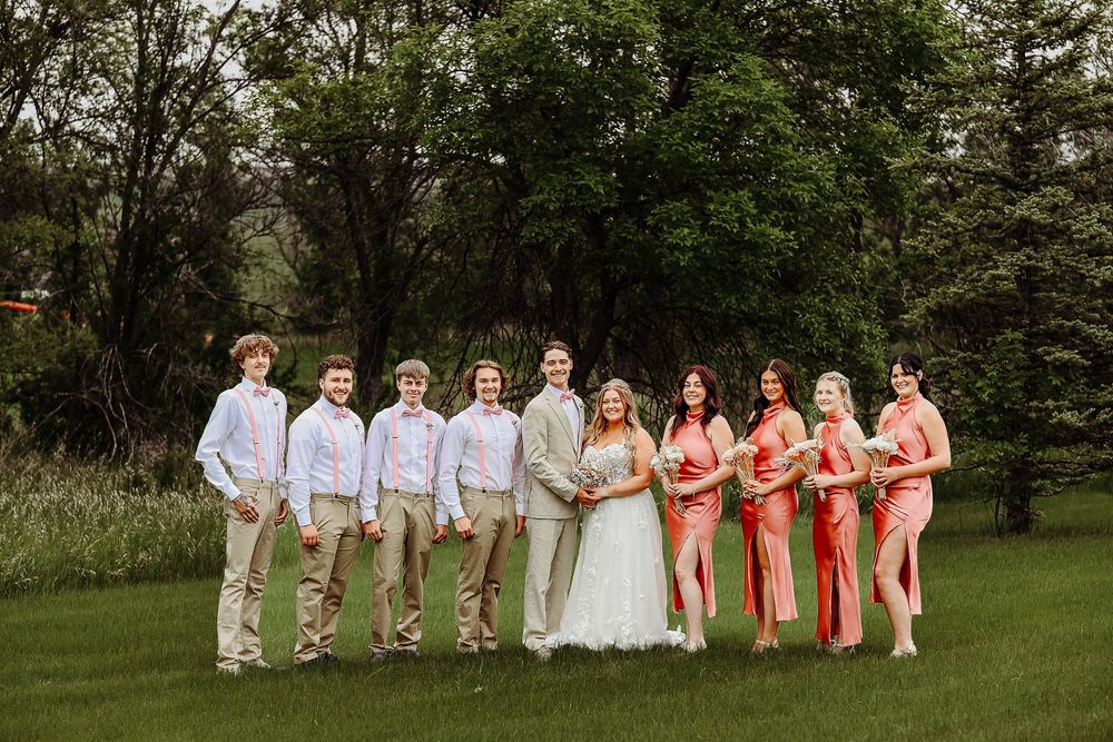 Wedding party in coral dresses and khaki suits poses together on lush green lawn at outdoor celebration.