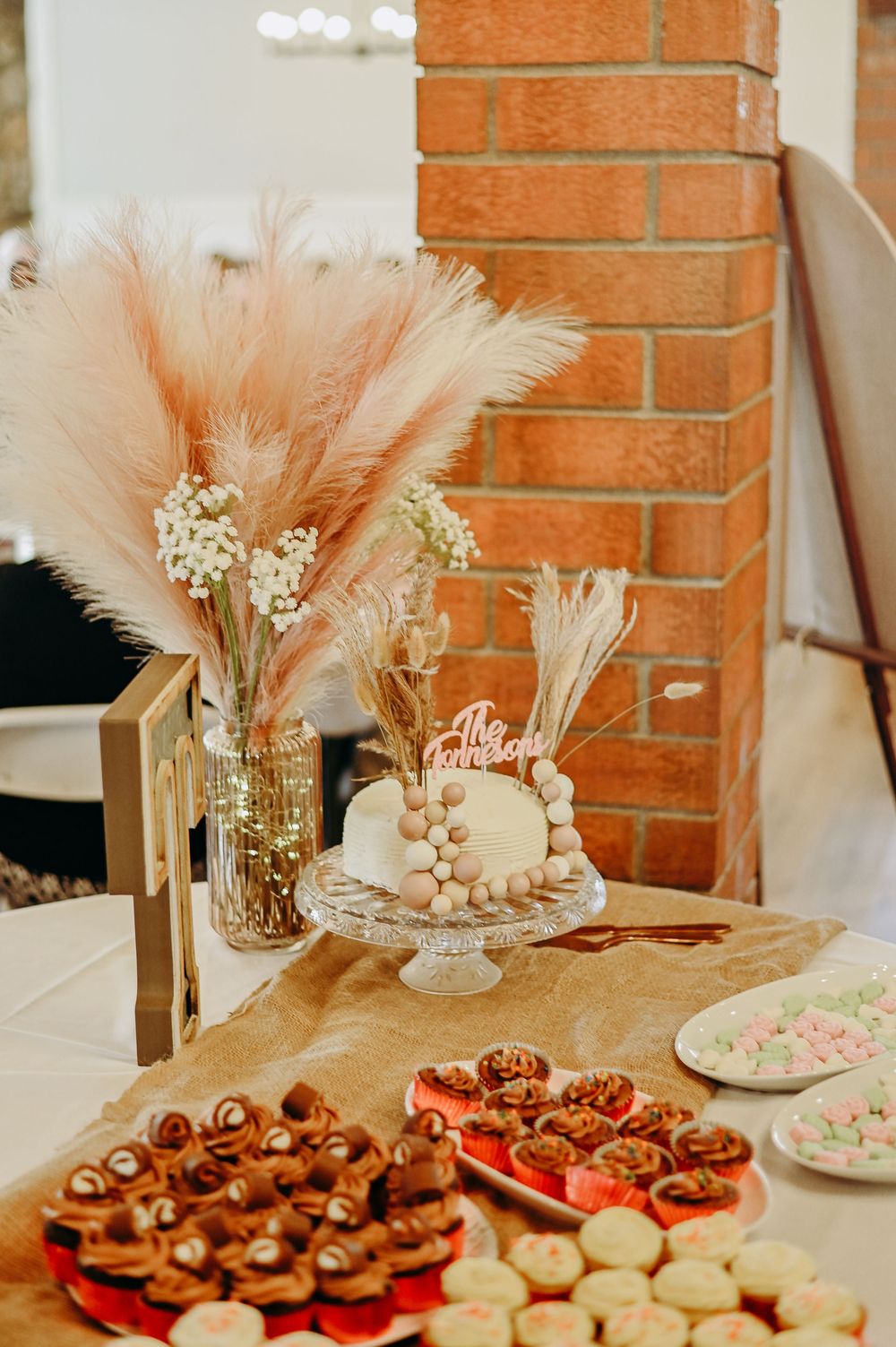 Elegant dessert table with chocolate cupcakes and pampas grass decorations against a rustic brick wall.
