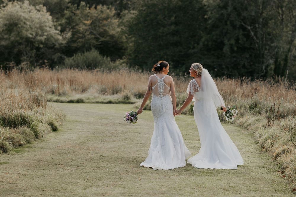 Two brides in white lace wedding dresses holding hands walking through golden meadow.