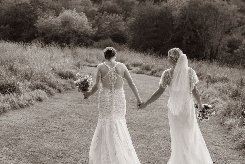 Black and white photo of two brides in lace gowns holding bouquets walking hand in hand.