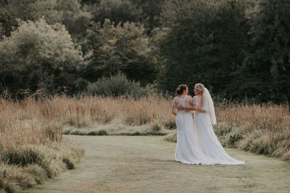 Two brides in elegant white wedding dresses standing together in a scenic meadow landscape.