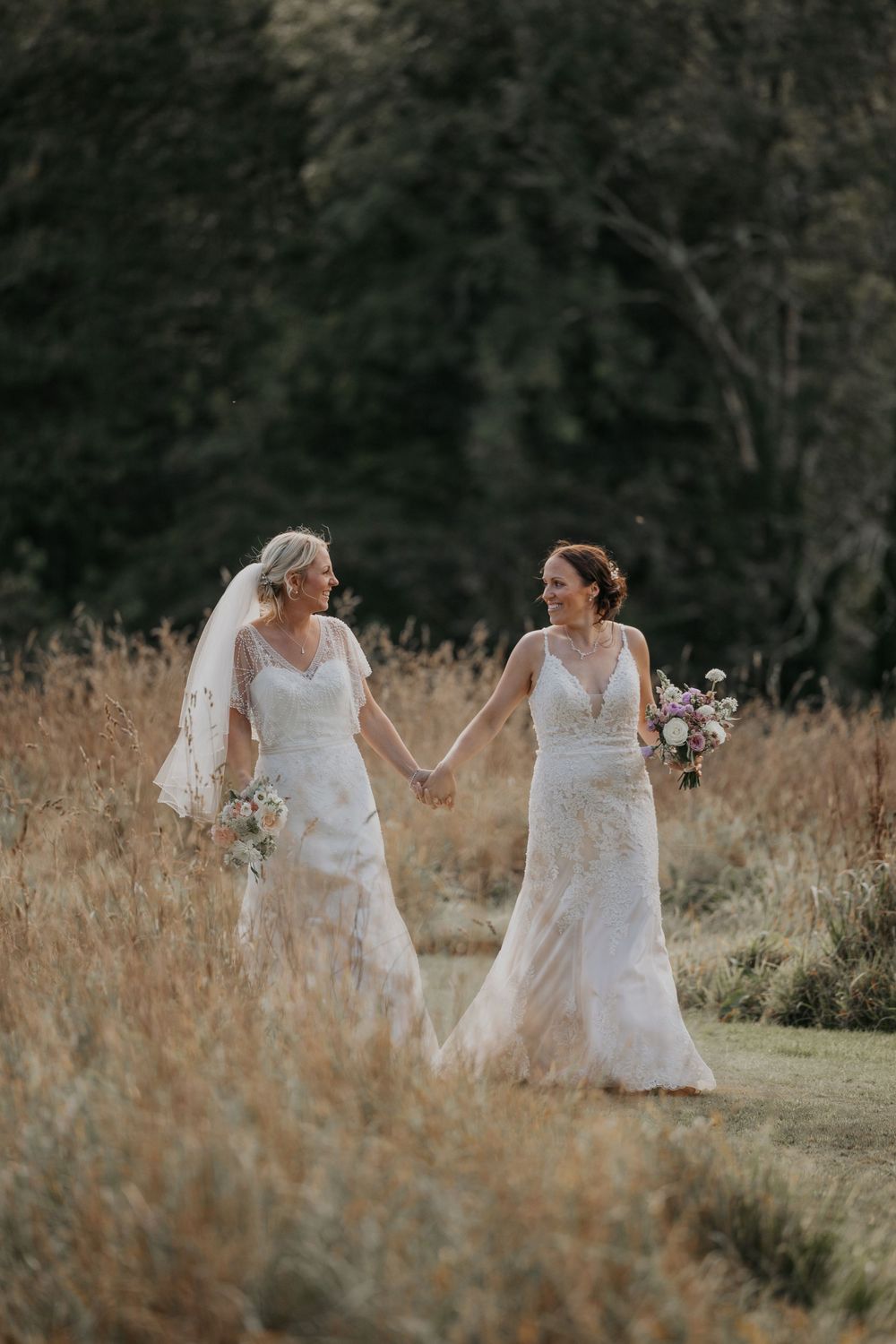 Two brides in beaded lace wedding gowns holding bouquets and smiling in golden field.