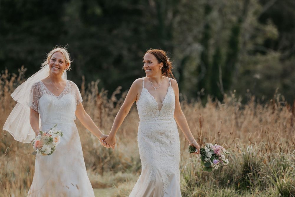 Two joyful brides in white lace wedding dresses holding hands and bouquets in sunny meadow.