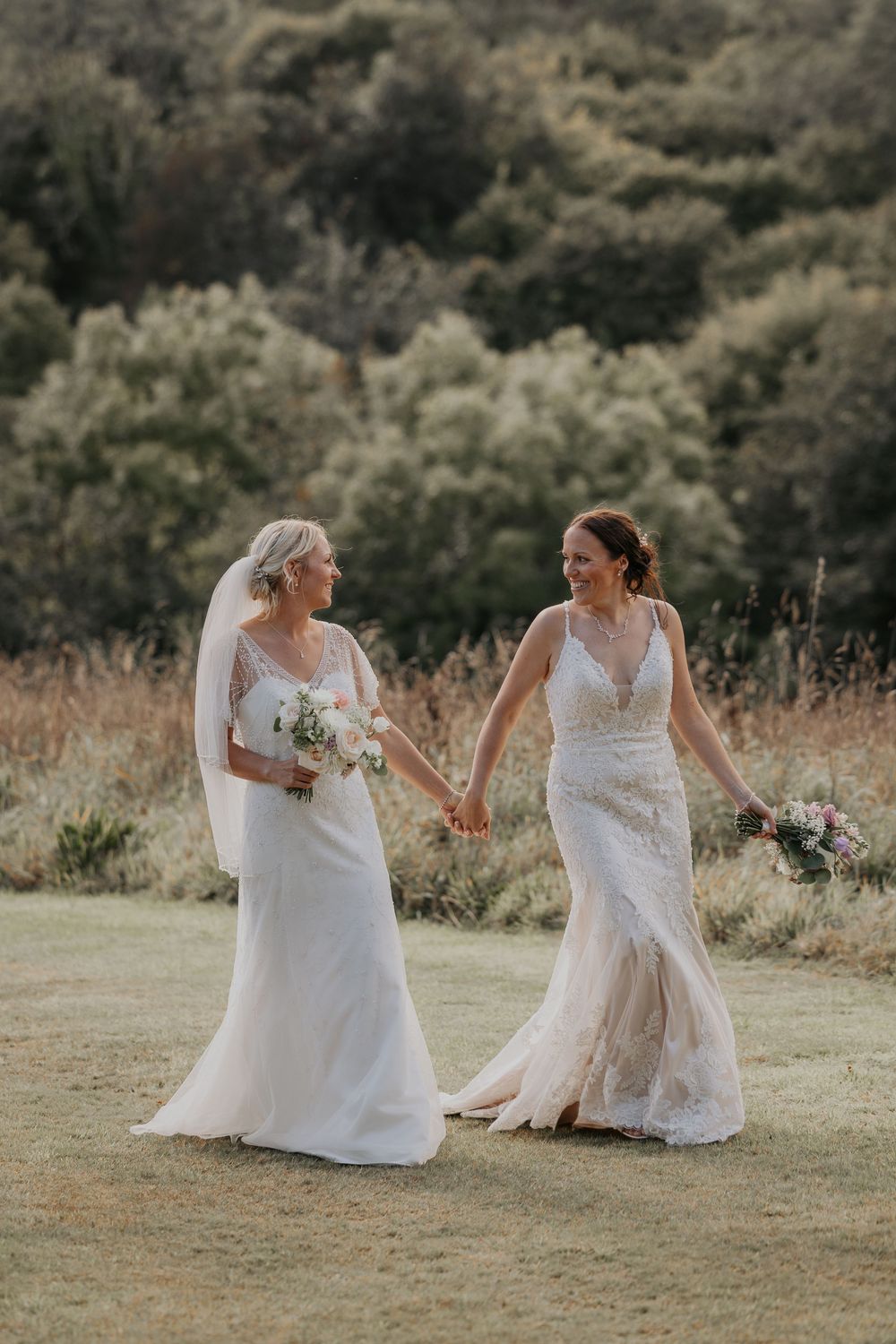 Two brides in white lace wedding dresses holding hands and bouquets in sunlit countryside.