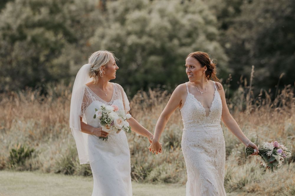 Two smiling brides in white lace wedding gowns holding bouquets walking together outdoors.
