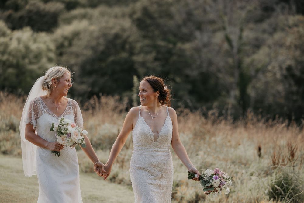 Two brides in white wedding gowns hold hands and smile while walking through a scenic outdoor meadow.