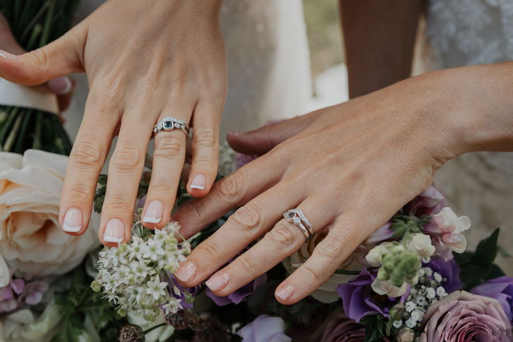 Two brides display their wedding rings over a bouquet of white and purple flowers.
