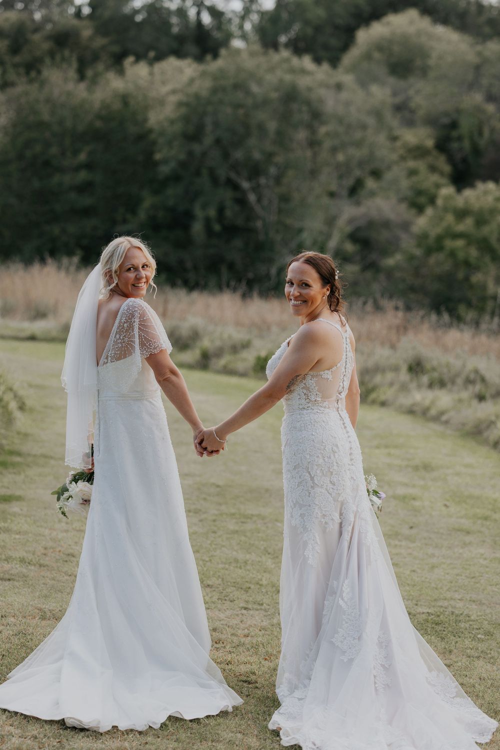 Two brides in elegant white lace wedding gowns hold hands and smile on a lush green lawn.