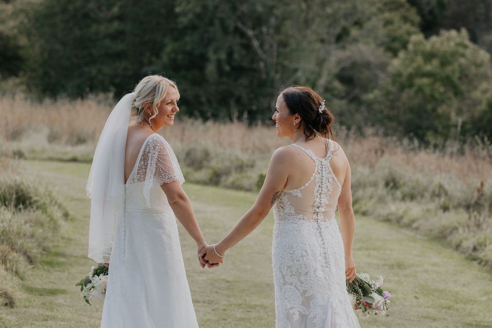 Two brides in white lace wedding dresses hold hands while walking along a scenic outdoor path.