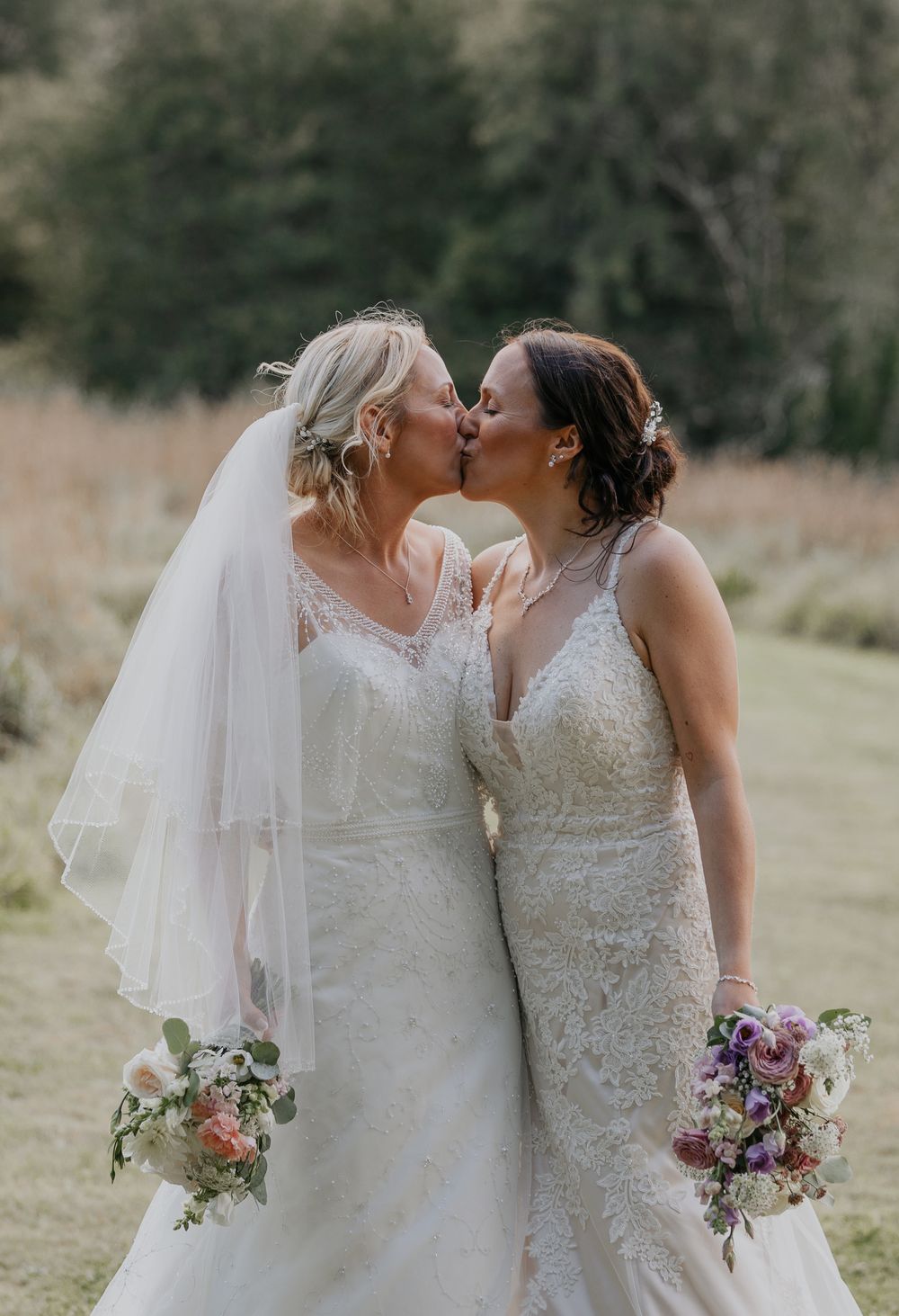 Two brides in white lace wedding gowns share a kiss while holding colorful bridal bouquets outdoors.