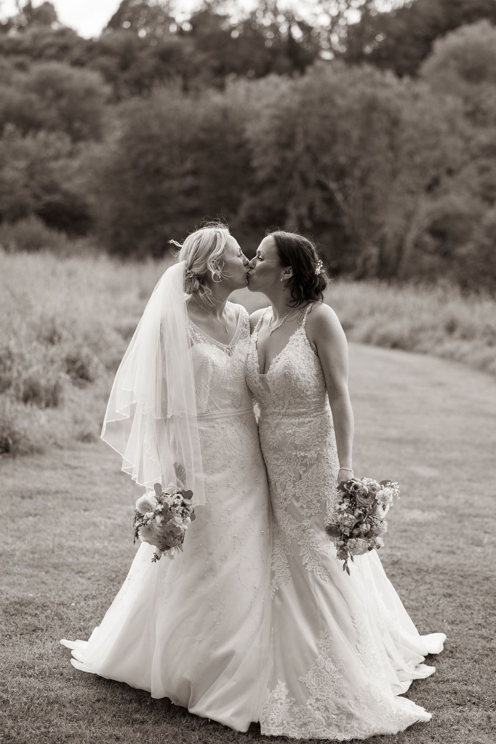 Black and white photo of two brides kissing while holding bouquets in an outdoor garden setting.