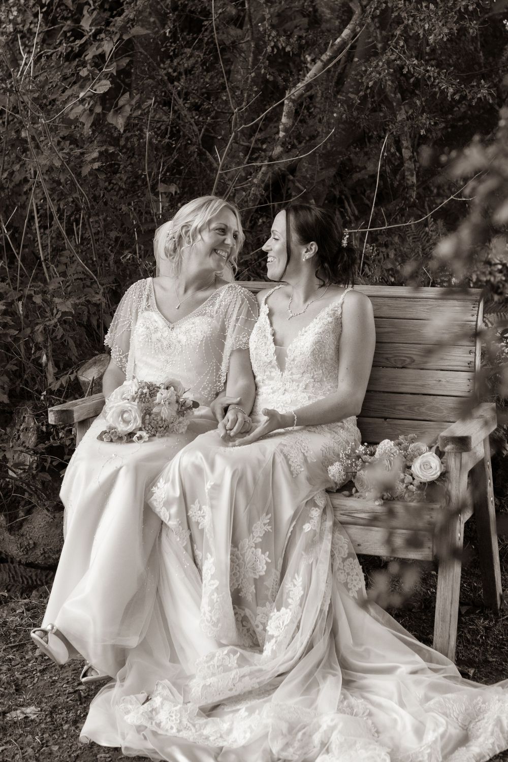 Black and white photo of two brides sitting on a wooden bench, smiling at each other.