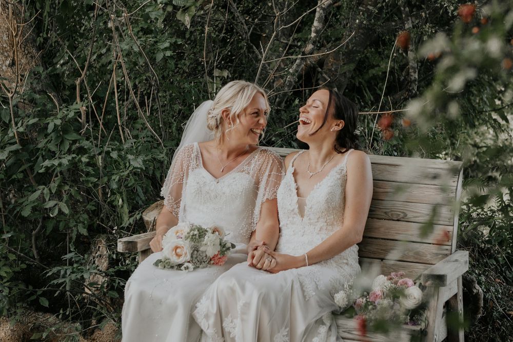 Two brides in white lace wedding gowns laugh joyfully while sitting on a rustic wooden bench outdoors.