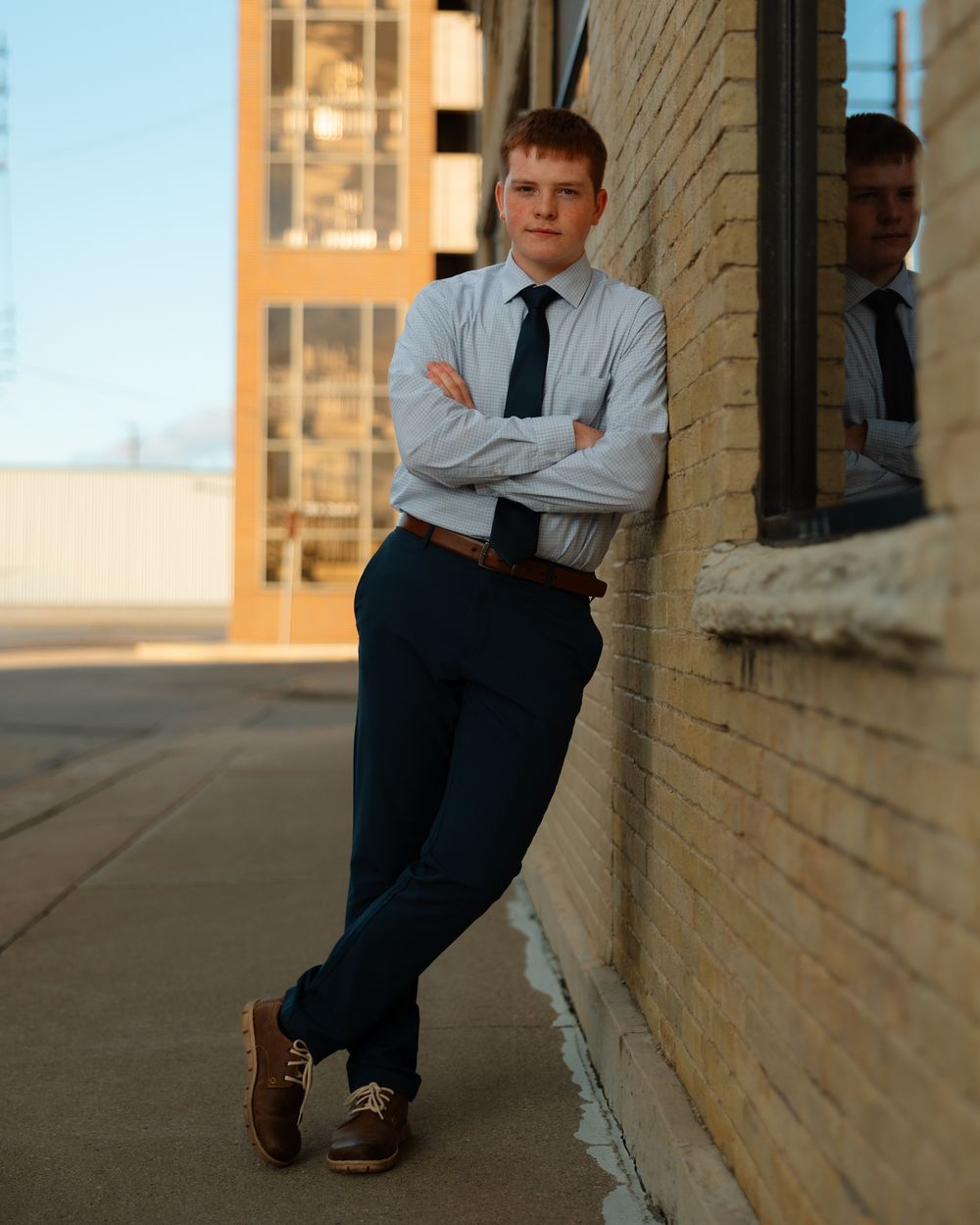 A person in business attire leans against a brick wall at sunset, wearing a light blue shirt and dark tie.