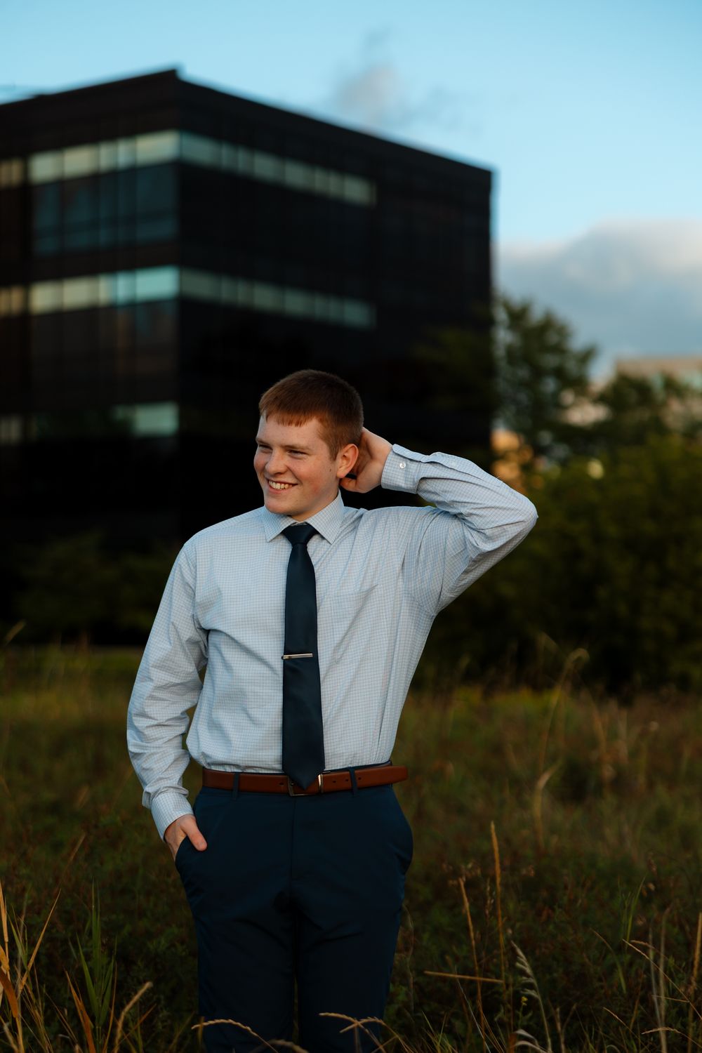 A person in business attire poses in a field at sunset with office buildings visible in the background.