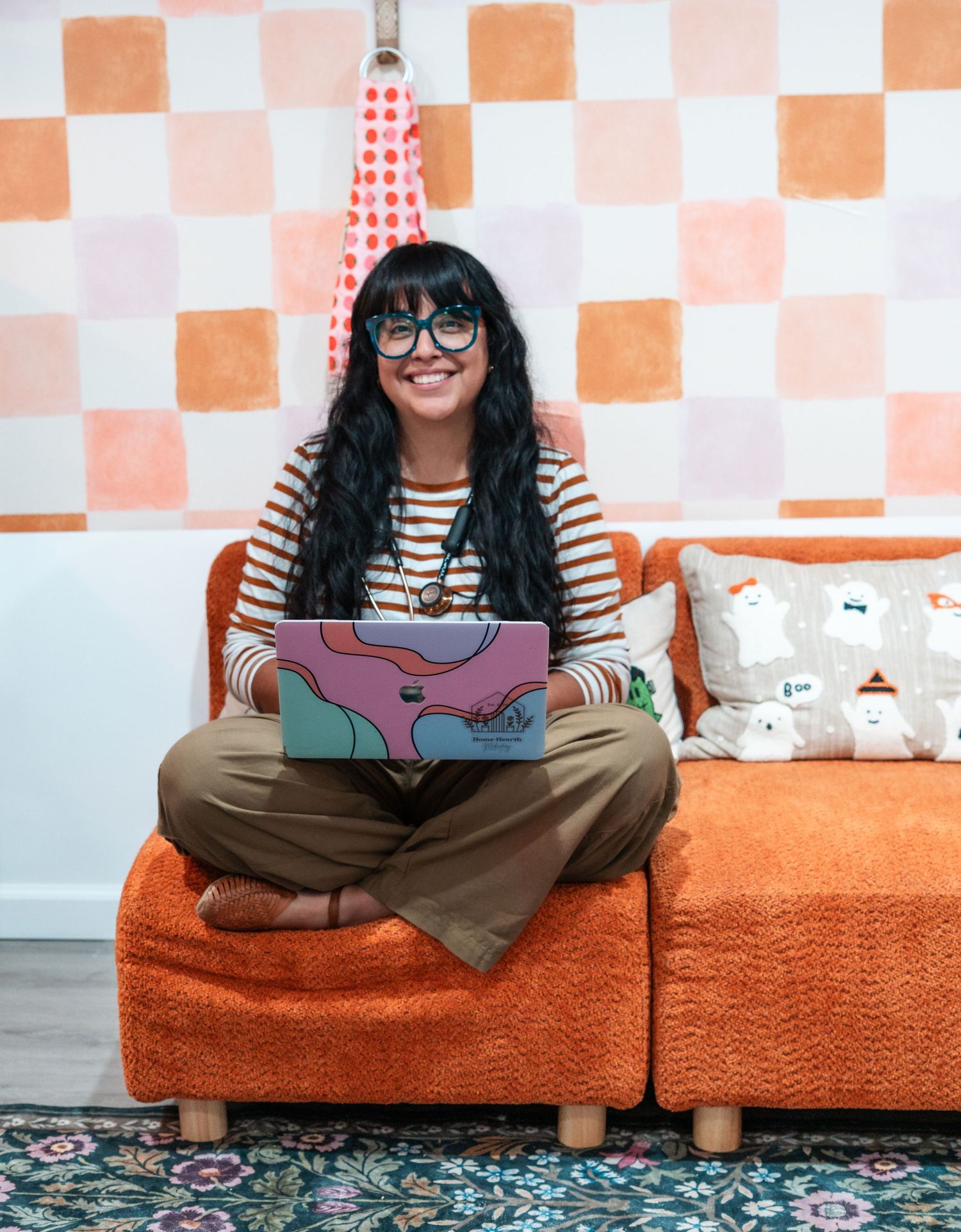 A person in glasses and stripes sits cross-legged on an orange couch with a patterned rug below.