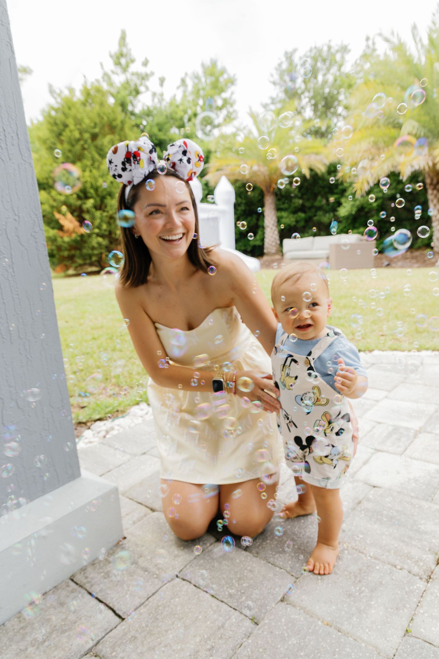 A smiling mother wearing Mickey Mouse ears kneels beside her baby boy as colorful bubbles float around them outdoors.