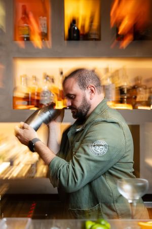 A person in a green jacket uses a cocktail shaker while working behind a dimly lit bar counter.