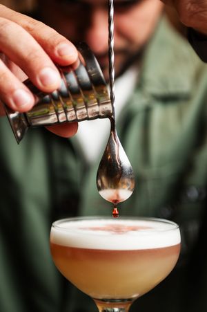 A bartender pours a craft cocktail from a mixing tin into a coupe glass, creating a frothy pink drink.