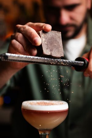 Bartender grating ice over a pink cocktail in a coupe glass, creating a snowy garnish effect.