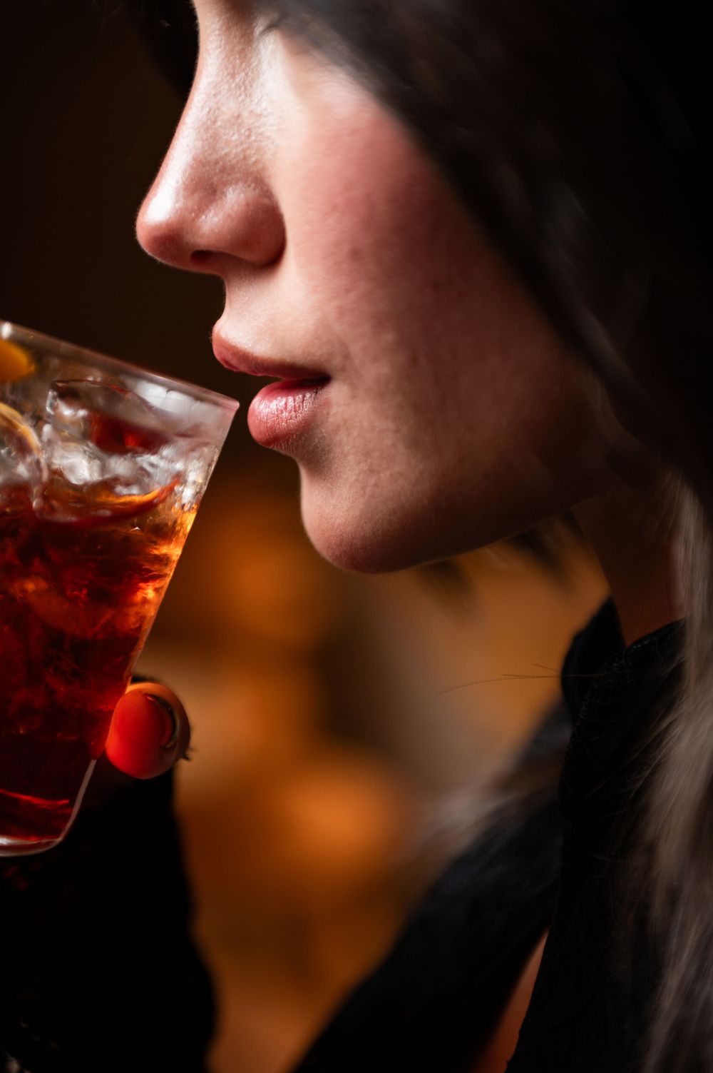 A person sips a red cocktail with a cherry garnish in a dramatic side profile silhouette against warm lighting.