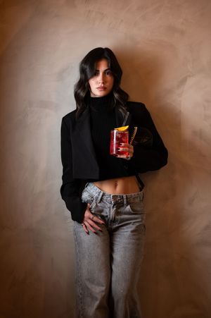 Woman in black cropped blazer and turtleneck with gray jeans holds a decorative cup against a beige wall.