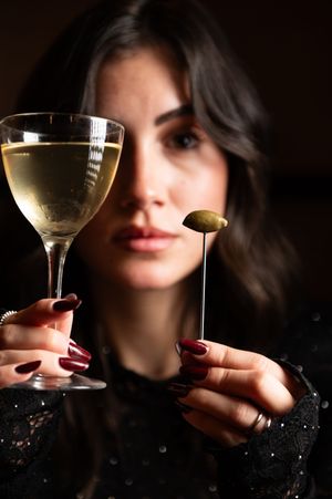 Woman holding a martini glass and olive garnish on a cocktail pick against a dark background.
