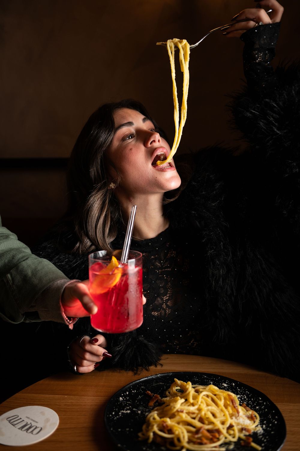 Person eating spaghetti and drinking red cocktail in dramatic lighting at restaurant table.