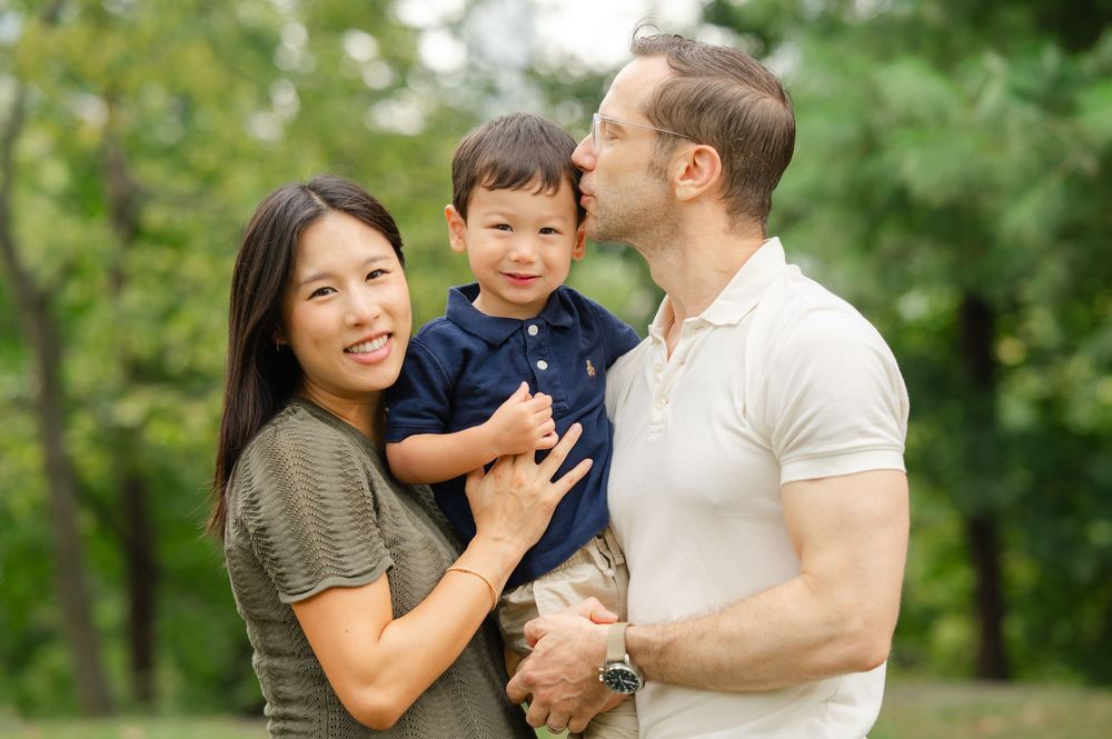 A family enjoying a playful moment together outdoors in a park with green trees in the background.