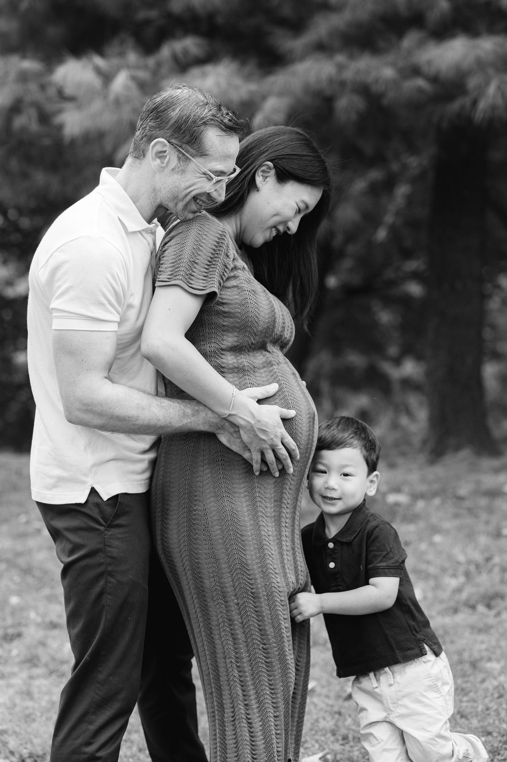 A touching black and white photo of a couple with their son leaning against his mother's pregnant belly while the father embraces her.
