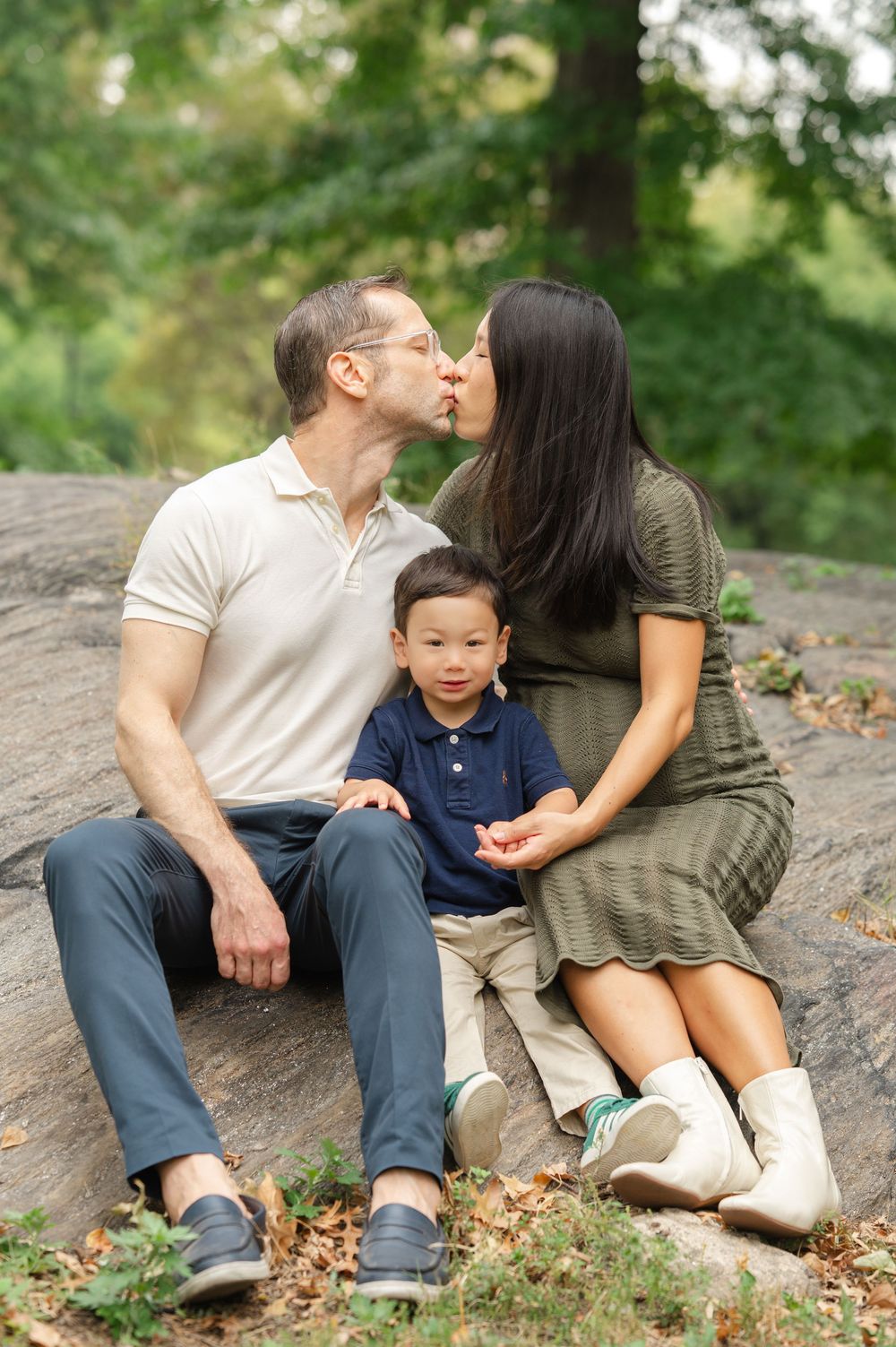 A family sits together on a large rock surrounded by fallen autumn leaves in a park setting.