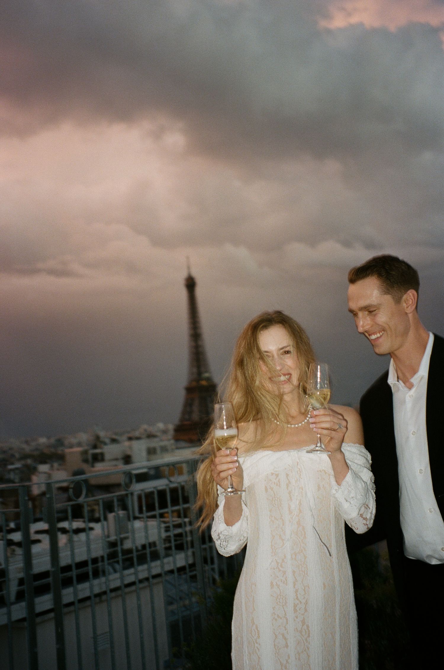 A couple shares champagne on a rooftop terrace in Paris with the Eiffel Tower and moody sunset sky in the background.