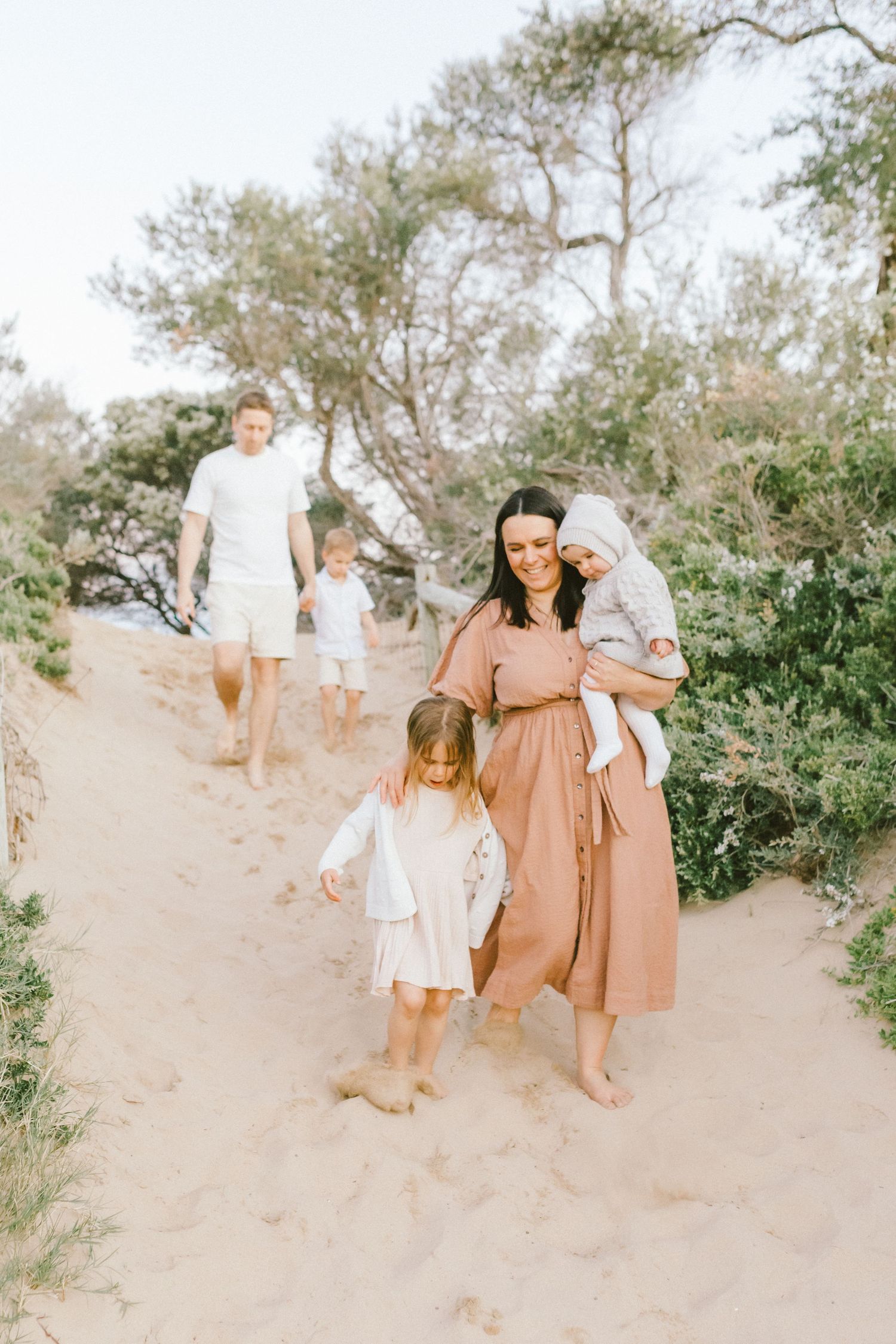 Family walking together on sandy path through desert landscape wearing neutral colored clothing.