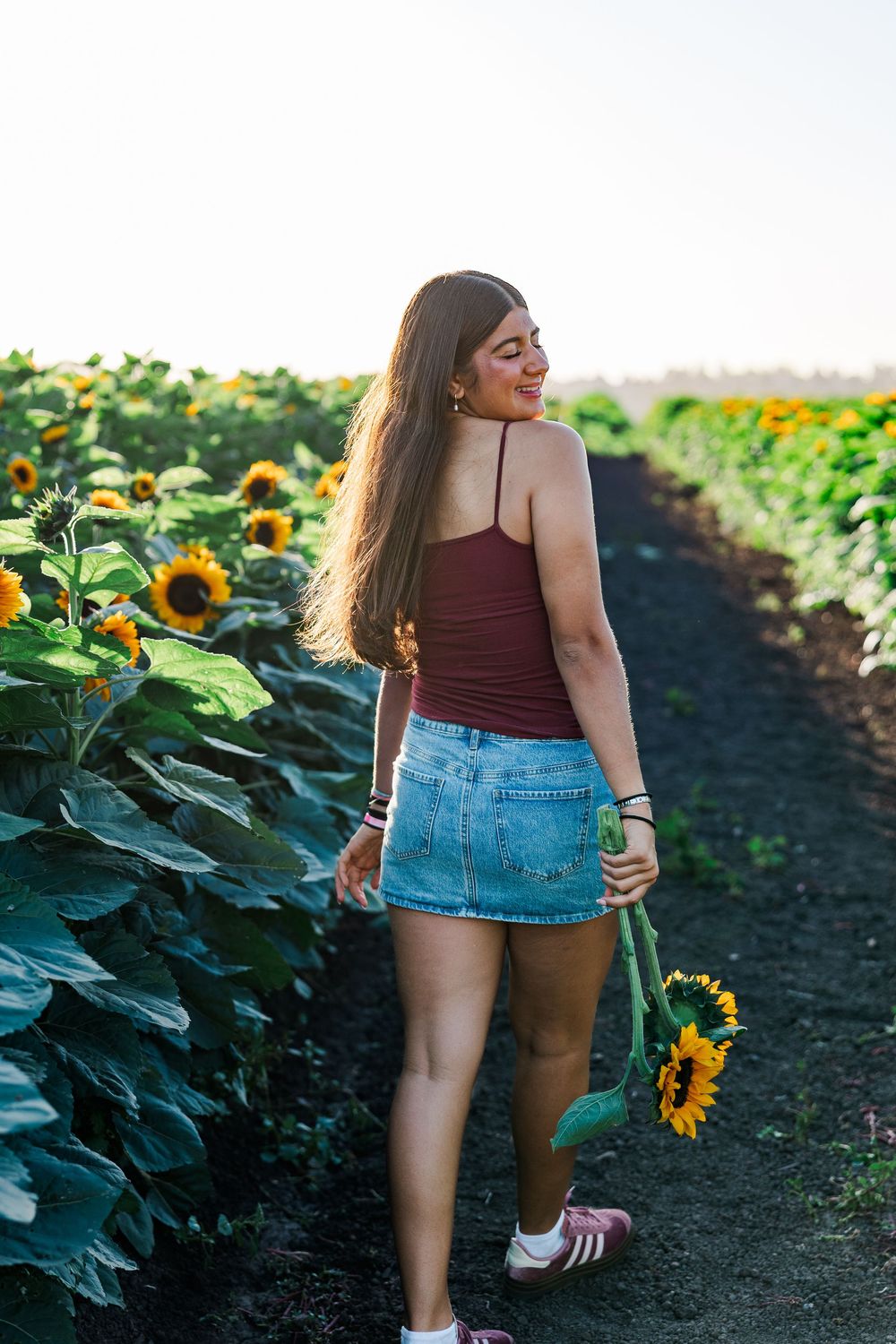 Woman in burgundy top and denim shorts walking through vibrant sunflower field during golden hour.